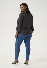 Blouse noire texturée avec des manches trois-quarts, associée à un jean skinny bleu et des sandales à talons hauts noires. Fond clair et léger.