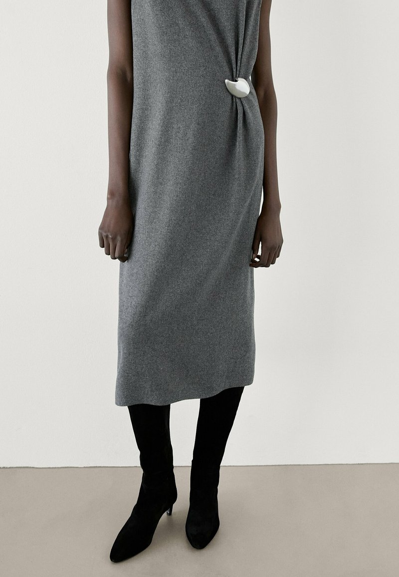 Robe midi grise sans manches avec un détail drapé sur le côté fixé par une boucle blanche, portée avec des bottes à talons noires sur un fond neutre.