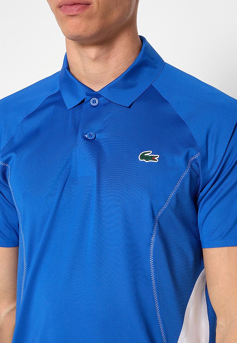 Polo shirt bleu avec un col classique, des manches courtes et un logo de crocodile vert sur la poitrine gauche. Tissu lisse avec des accents texturés.