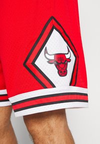 Czerwone sportowe szorty z naszywką z logo Chicago Bulls oraz czarno-czerwono-białym paskiem na brzegu, noszone przez osobę.