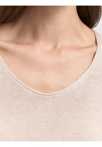 Haut en tricot beige clair avec un col en V, mettant en valeur une texture lisse et des coutures discrètes le long de l'encolure. Vue en gros plan du tissu.