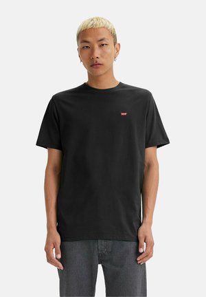 Mand med kort blond hår iført en sort Levi's t-shirt og grå jeans, stående mod en ensfarvet hvid baggrund.