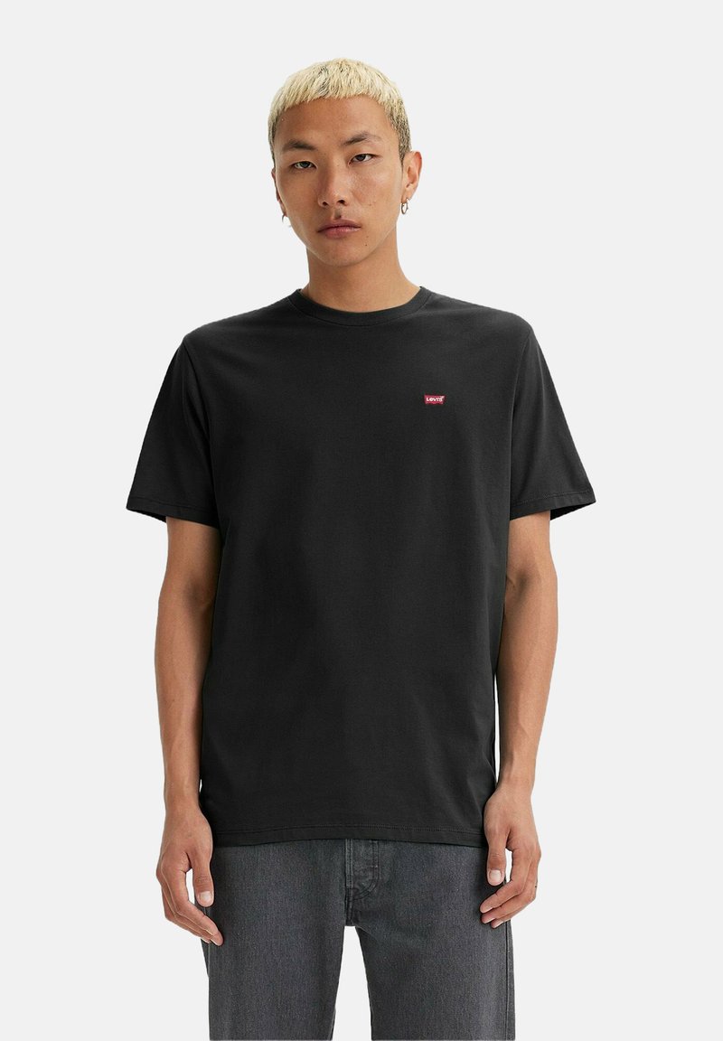 Uomo con capelli biondi corti vestito con una t-shirt nera Levi's e jeans grigi, in piedi contro uno sfondo bianco semplice.