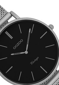 Montre à cadran noir avec des index et aiguilles argentés. Équipée d'un bracelet en acier inoxydable tissé et d'un design minimaliste. Marquée "OOZOO Vintage."