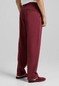 Pantaloni bordeaux dal taglio rilassato, con una tasca posteriore con bottone, abbinati a pantofole bordeaux. Tessuto dalla texture liscia.