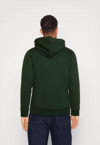 Felpa con cappuccio verde scuro, dotata di un cappuccio ampio, polsini e orlo a costine, realizzata in un tessuto morbido e strutturato. Indossata con jeans in denim scuro.