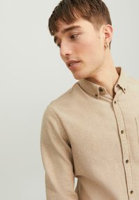 Beige Hemd mit Knopfleiste aus strukturiertem Stoff, mit Kragen, Fronttasche und braunen Knöpfen. Modell ist im Seitenprofil abgebildet.