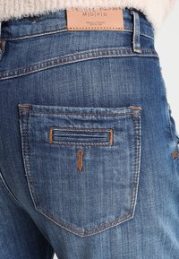 Siniset farkut, joissa on nahkainen vastaus, jossa lukee "Marc O'Polo Denim." Takataskuissa on koristeompeleet ja ne ovat sileät.