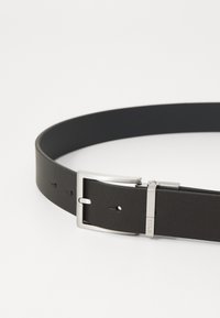 Ceinture en cuir noir avec une texture lisse et une boucle en métal argenté, dotée de plusieurs trous pour l'ajustement et d'un design élégant.