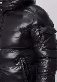 Doudoune noire à finition brillante, dotée d'une fermeture éclair sur le devant, de poches latérales et d'une capuche amovible. Agrémentée de détails de couture.