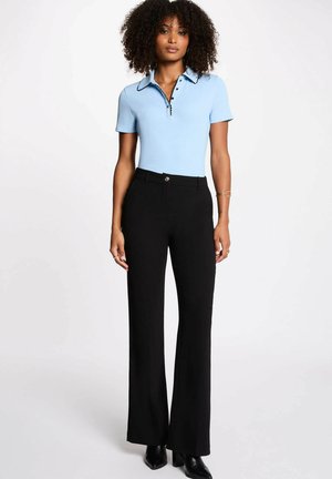 Donna con capelli ricci che indossa una maglietta azzurro chiaro a maniche corte con colletto e pantaloni larghi a vita alta neri, abbinati a stivali neri con tacco.