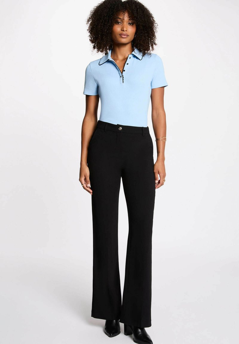 Femme aux cheveux bouclés portant un haut à manches courtes bleu clair avec col et un pantalon noir taille haute à jambes larges, accompagné de bottes à talons noires.