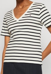 T-shirt a righe con scollo a V in crema e blu navy. Maniche corte, design aderente, con un piccolo logo sul lato superiore sinistro. Tessuto morbido.