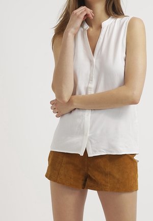 Kvinde iført en hvid, ærmeløs skjorte med knapper og brune ruskinds-shorts, med den ene arm krydset og den anden hånd tæt på hagen.