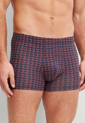 Torso e parte superiore delle gambe maschili che indossano boxer scuri con un motivo ricorrente di linee curve rosse e arancioni su uno sfondo grigio chiaro.