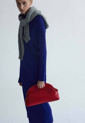 Femme portant une longue robe bleue et un pull gris posé sur les épaules, tenant une grande pochette rouge sur un fond uni.
