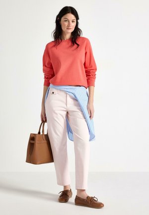 Donna che indossa una felpa color corallo, pantaloni rosa chiaro, camicia a righe blu annodata in vita, mocassini marroni e che porta una borsa marrone, in piedi su sfondo bianco.