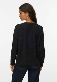 Blusa negra de manga larga con patrones geométricos texturizados, escote redondeado y corte relajado. La parte de atrás tiene un detalle de fruncido sutil.
