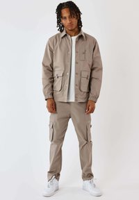 Giacca leggera beige con quattro tasche, chiusura con bottoni e polsini elastici, abbinata a pantaloni cargo coordinati. Sneakers bianche completano il look.