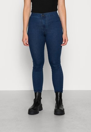 Person iført mørkeblå skinny jeans, sort ribbet ærmeløs top og sorte chunky ankelstøvler, stående på en lys grå gulv mod en hvid væg.