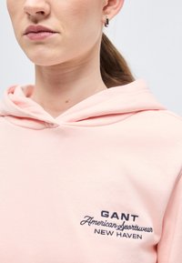 Približen posnetek osebe, ki nosi svetlo roza kapucar z vezenim napisom "GANT American Sportswear NEW HAVEN" na prsih.