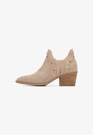 Stivaletto alla caviglia in camoscio beige con punta affusolata, dettagli decorativi con borchie sui lati e tacco impilato in legno.