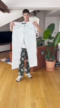 Šviesiai mėlyni džinsai, laikomi rankoje, su penkių kišenių dizainu, diržo kilpomis ir matomais siūlais. Modelis vilki raštuotas žalias kelnes ir sportbačius.
