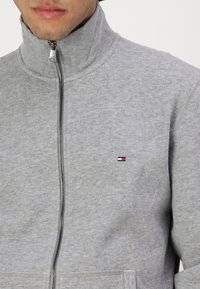Grauer Kapuzenpullover aus weichem Stoff, mit hohem Kragen und einem kleinen, in Rot, Weiß und Blau auf der Brust gestickten Logo.