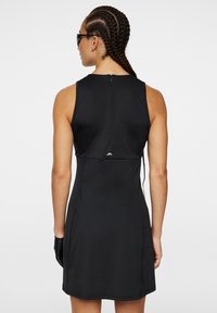 Robe noire sans manches en tissu lisse, avec un col haut, une fermeture éclair au dos, et un accent logo discret près de l'ourlet.