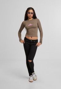 Barna, hosszú ujjú felső rózsaszín felirattal, fekete, szakadt farmernadrággal és fehér sportcipővel párosítva. A modellen hosszú, hullámos haj és napszemüveg van.