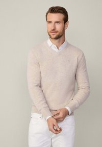 Jasno beżowy sweter dzianinowy z dekoltem w serek, z prążkowanymi mankietami i dołem, noszony na białej koszuli z kołnierzykiem, w połączeniu z białymi spodniami.