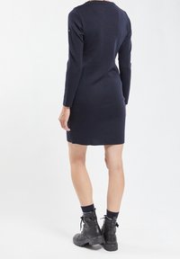 Personne portant une robe noire à manches longues, longueur genou, avec des bottines noires, debout de dos sur un fond blanc.