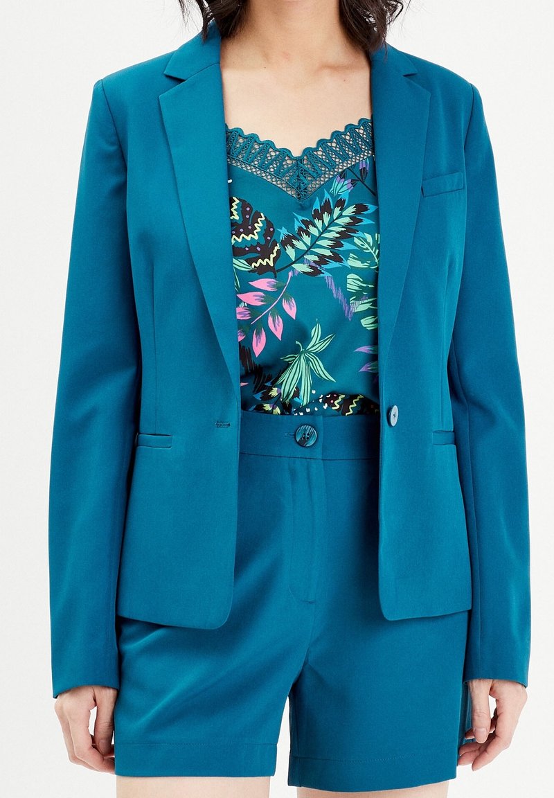 Blazer bleu sarcelle et short taille haute assorti portés sur un caraco en dentelle florale bleu sarcelle, visibles des épaules jusqu'à mi-cuisse.