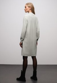 Lichtgrijze gebreide jurk met lange mouwen, tot halverwege de dijen, met een lichte textuur, gecombineerd met zwarte enkellaarsjes. Eenvoudig, modern ontwerp zonder patronen.