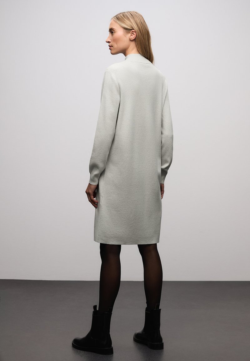 Lichtgrijze gebreide jurk met lange mouwen, tot halverwege de dijen, met een lichte textuur, gecombineerd met zwarte enkellaarsjes. Eenvoudig, modern ontwerp zonder patronen.