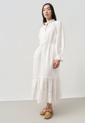 Vrouw in lange witte jurk met geborduurde zoom en lange mouwen, draagt beige gevlochten Mary Jane schoenen, staand tegen een effen achtergrond.