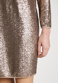 Robe en sequins bronze à manches longues, avec une texture scintillante et un ourlet arrondi. Les sequins créent un effet dégradé.