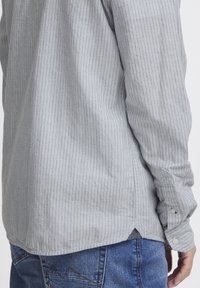 Camicia a righe grigia realizzata in tessuto leggero con una vestibilità ampia, caratterizzata da polsini con bottoni e un orlo laterale con spacco e dettagli di cucitura bianca.