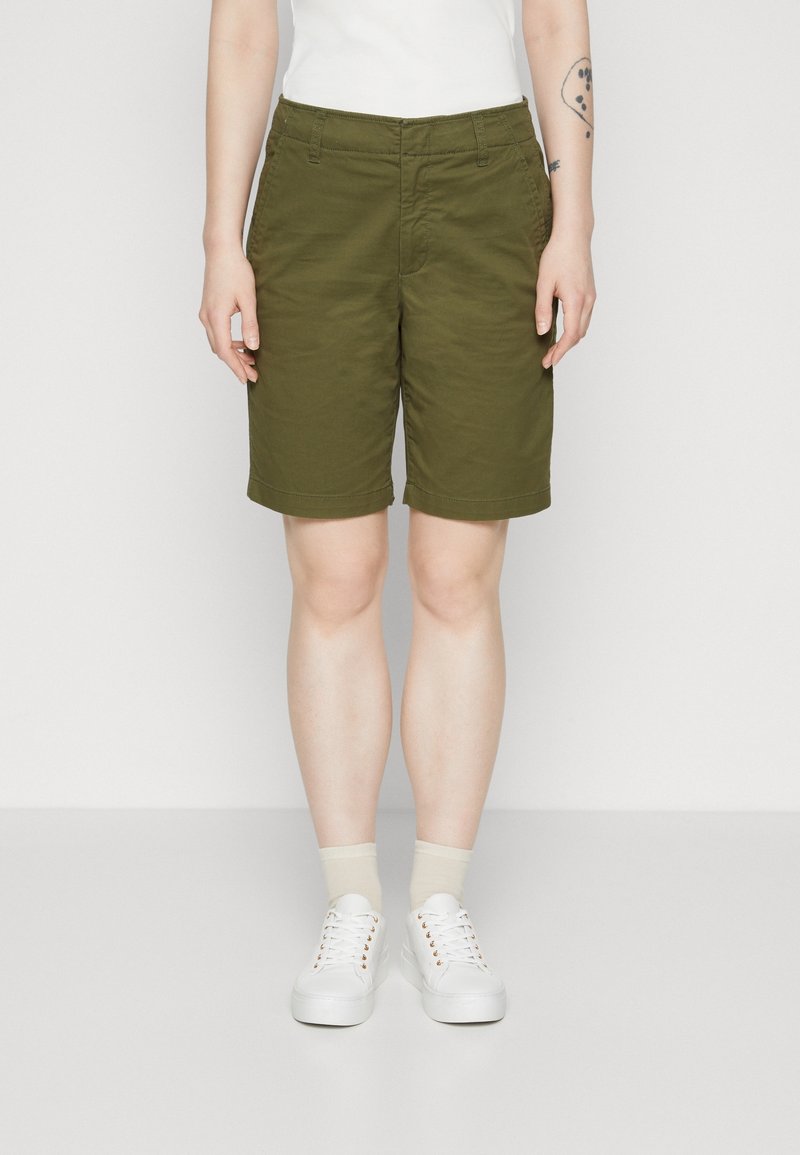 Shorts en coton vert olive à coupe droite, poches latérales apparentes et ourlets retroussés, associés à des baskets blanches et des chaussettes beige courtes.