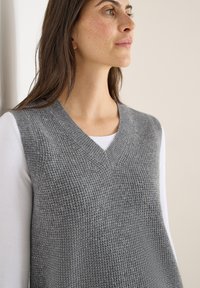 Maglione grigio senza maniche, lavorato a maglia, con scollo a V e motivo testurizzato, indossato sopra una camicia bianca a maniche lunghe. Tessuto morbido e vestibilità comoda.