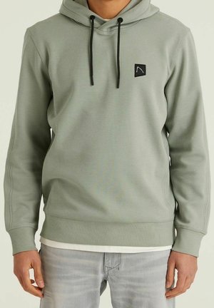 Mann trägt hellgrünen Hoodie mit schwarzen Kordeln und kleinem schwarzem Logo auf der Brust, kombiniert mit hellgrauen Jeans.