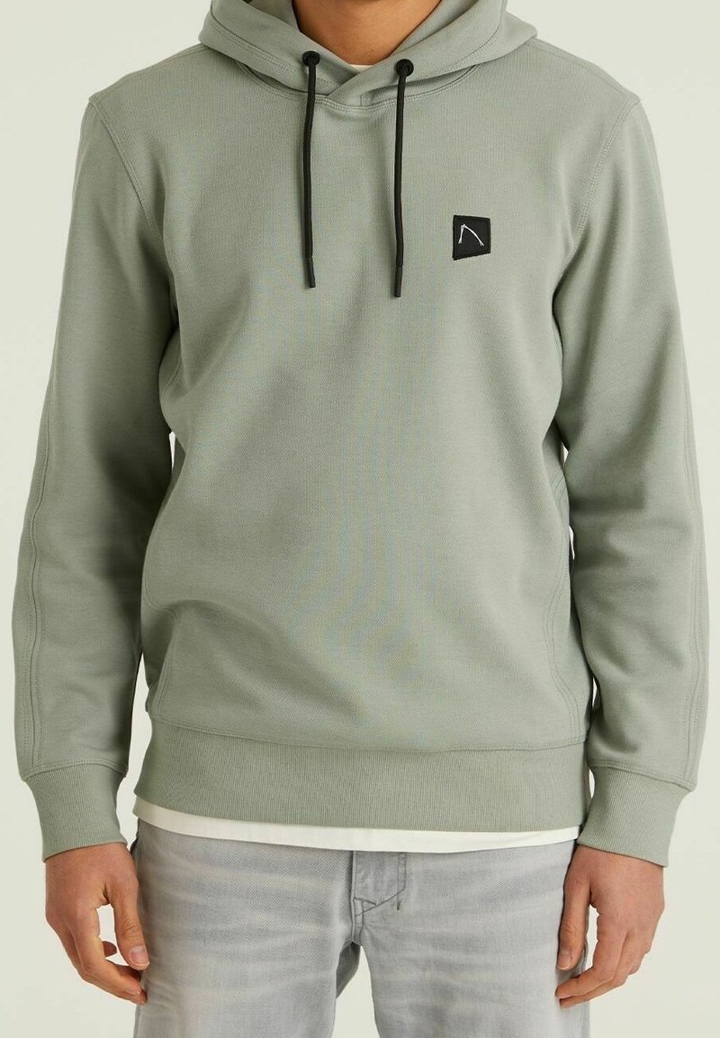 Homme portant un sweat à capuche vert clair avec des cordons noirs et un petit logo noir sur la poitrine, associé à un jean gris clair.