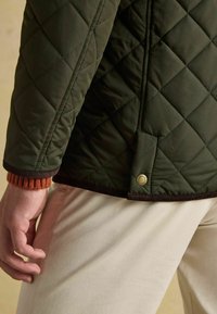 Person trägt eine dunkelgrüne gesteppte Jacke mit braunen Besätzen und beigen Hosen, zeigt Detail des Ärmels und des unteren Rückens mit geknöpftem Lüftungsschlitz.