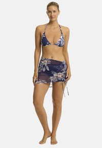 Modelo vistiendo un bikini floral azul marino y un pareo a juego, de pie descalza con un fondo neutro, sonriendo con confianza.