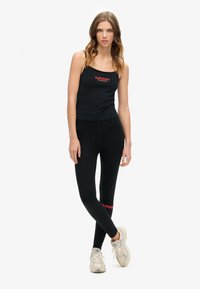 Canottiera nera con logo rosso "SUPERDRY", abbinata a leggings neri realizzati in tessuto morbido e elastico con un design minimale. Sneakers bianche completano il look.
