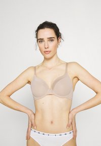 Beige polstret bh med justerbare stropper og en glatt tekstur. Har bøyle støtte og en rund design. Stilt med hvite truser.
