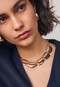 Gouden ketting met ovale schakels, gepolijst; gedragen bij een donkerblauw shirt. Simpel, modern ontwerp benadrukt de glans van de ketting.