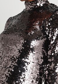 Multikolorierter Paillettenanzug mit hohem Kragen, der eine reflektierende Textur und eine taillierte Silhouette aufweist. Schimmert im Licht.