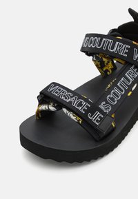 Mustad platvorm sandaalid laia, mustrilise rihmaga, millel on kirjas "Versace". Tekstuuriga sisetald, reguleeritav pandla ja julge disain.