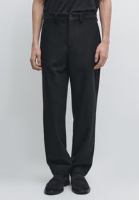 Pantalon noir à fines rayures avec une coupe structurée, un devant plat et des passants de ceinture, associé à des mocassins noirs sur un fond clair.